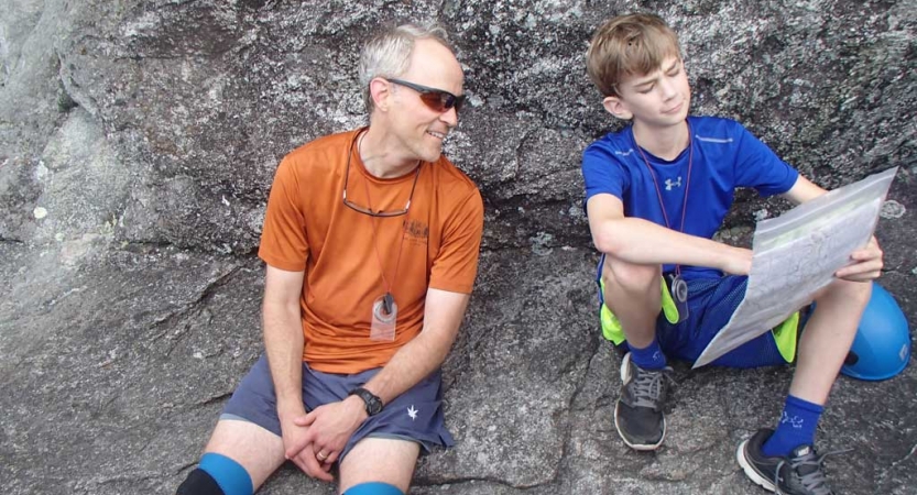 A parent and child rest on a rock and examine a map the child is holding and pointing too.